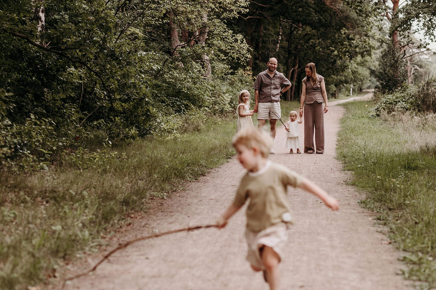 Jongen rent vooruit op een bospad tijdens een gezinsfotoshoot in 't Gooi, ouders en broertjes en zusjes lopen achter hem aan